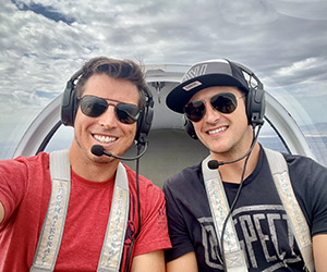 Two late 20's men wearing aviator sunglasses and communication headsets smiling in the cockpit of a plane in flight.